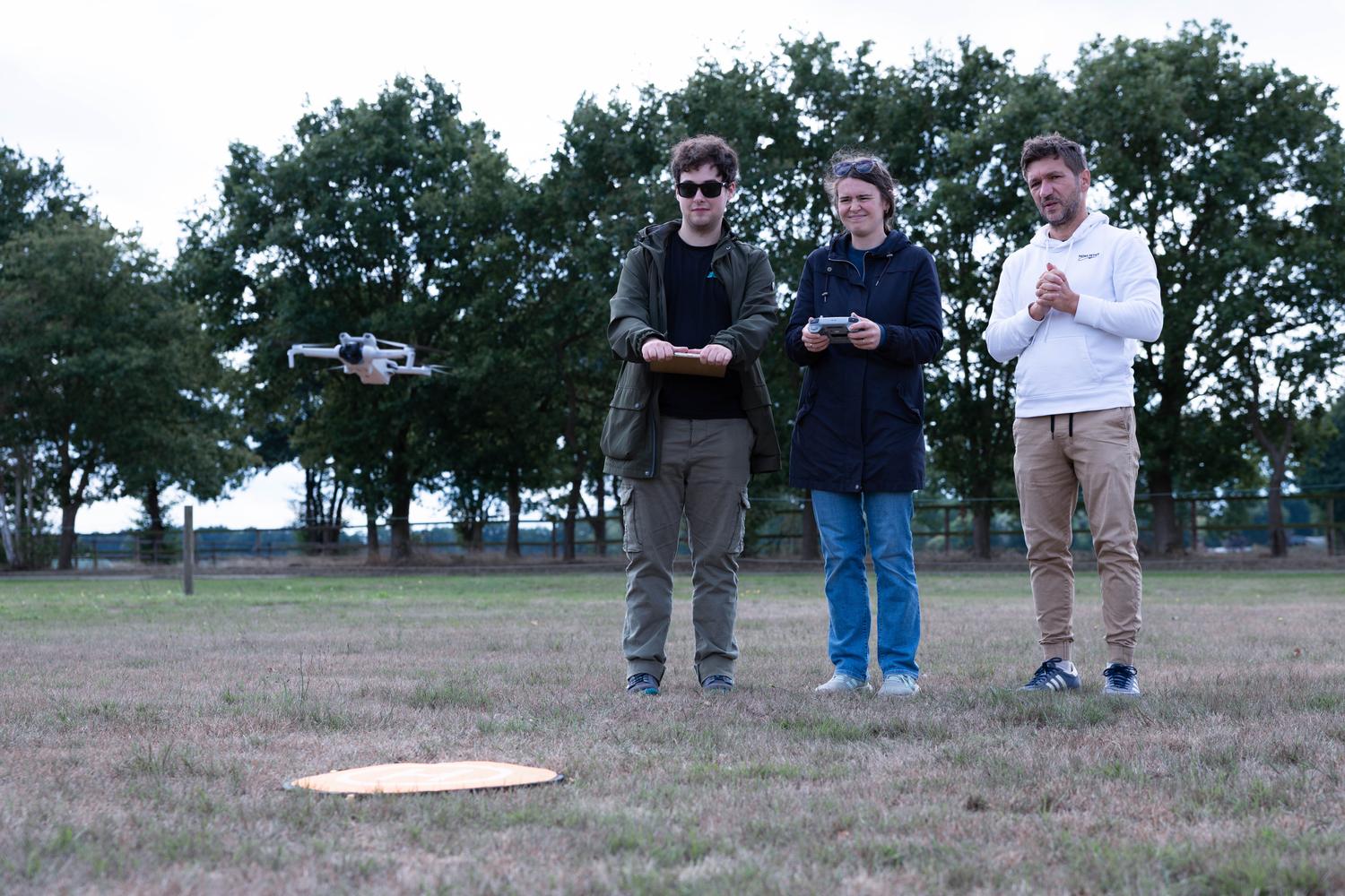 Drei Piloten mit Fernsteuerungen auf dem Flugfeld, links schwebt ihre Drohne über dem Landepad